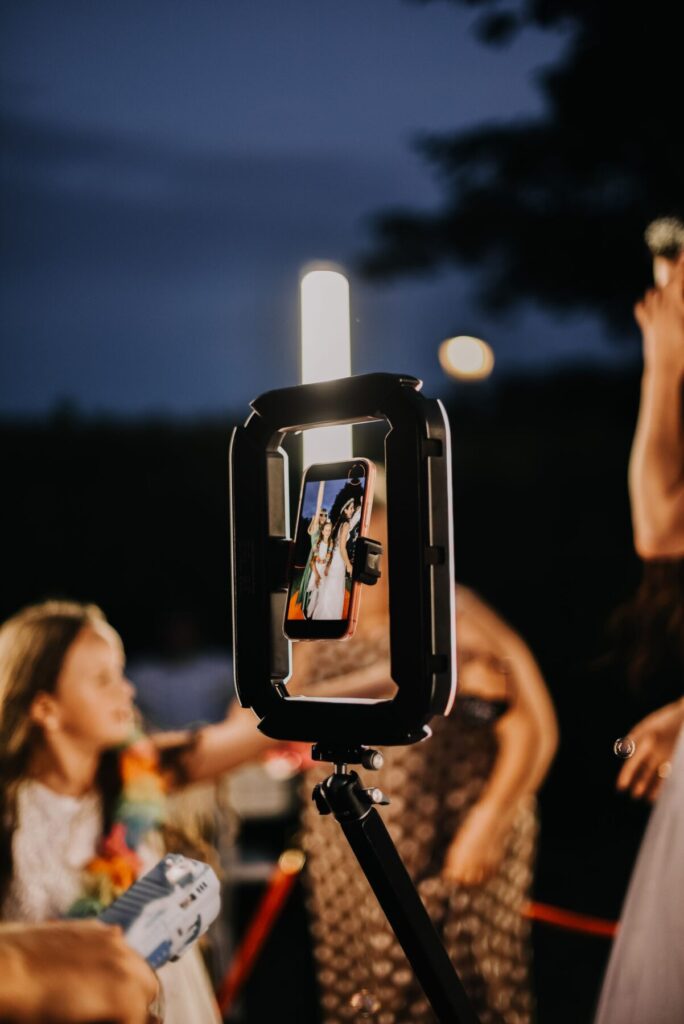 Fotobudka 360 w akcji, uchwycająca radosne chwile podczas imprezy. Na zdjęciu widoczny jest telefon umieszczony w fotobudce, rejestrujący uśmiechy gości na tle wieczornego klimatu. Idealne rozwiązanie na wesele lub przyjęcie, zapewniające wyjątkowe wspomnienia.